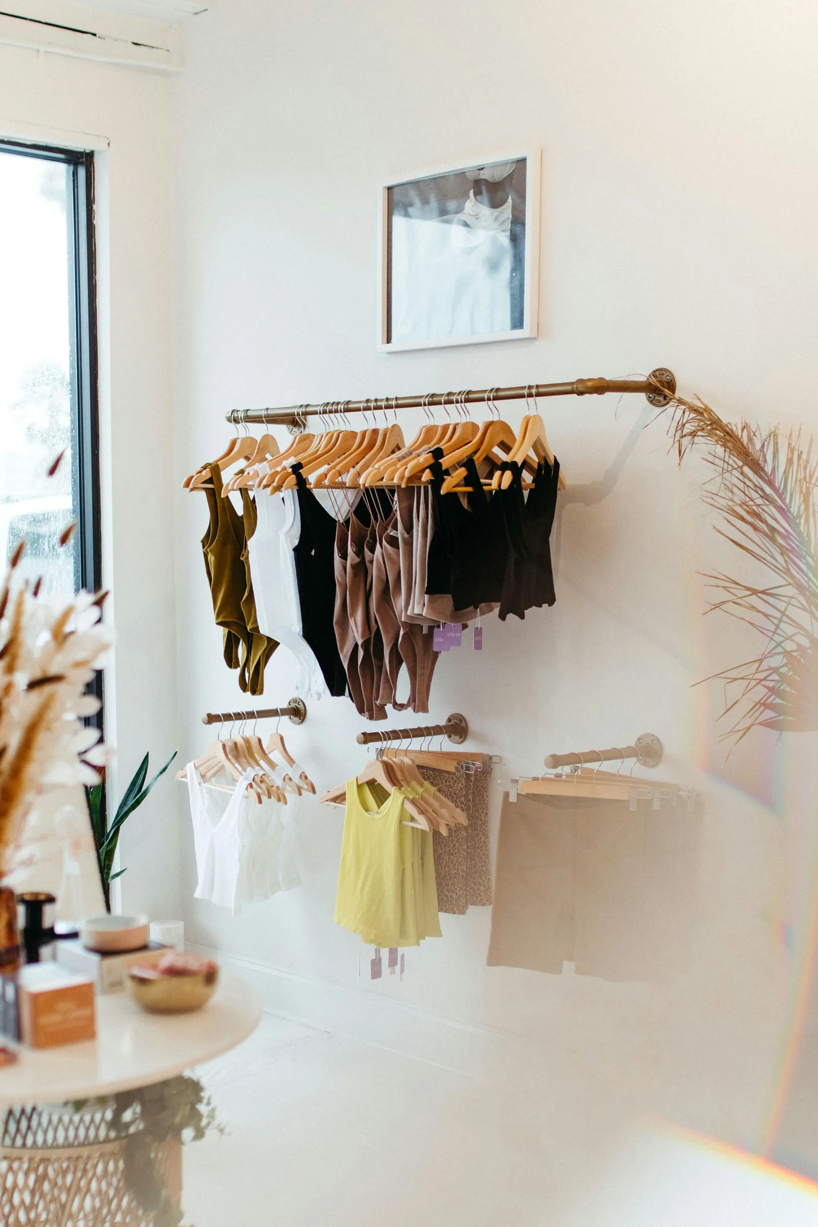 Boutique clothing display with tank tops hung on brass wall rods, framed art, and dried florals