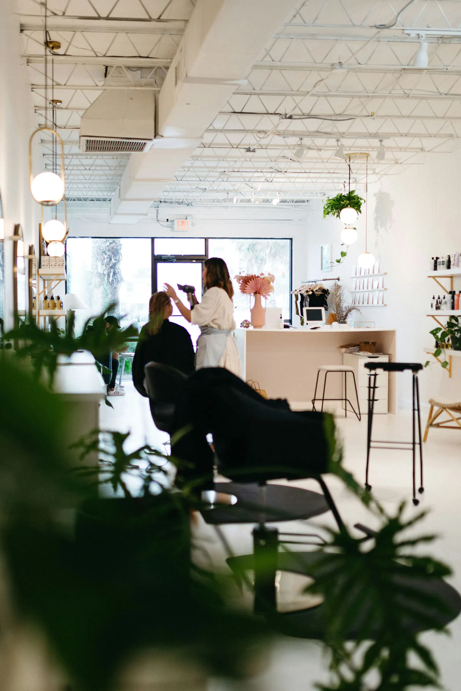 Stylist blow-drying a client's hair in a modern salon with greenery and an open industrial ceiling
