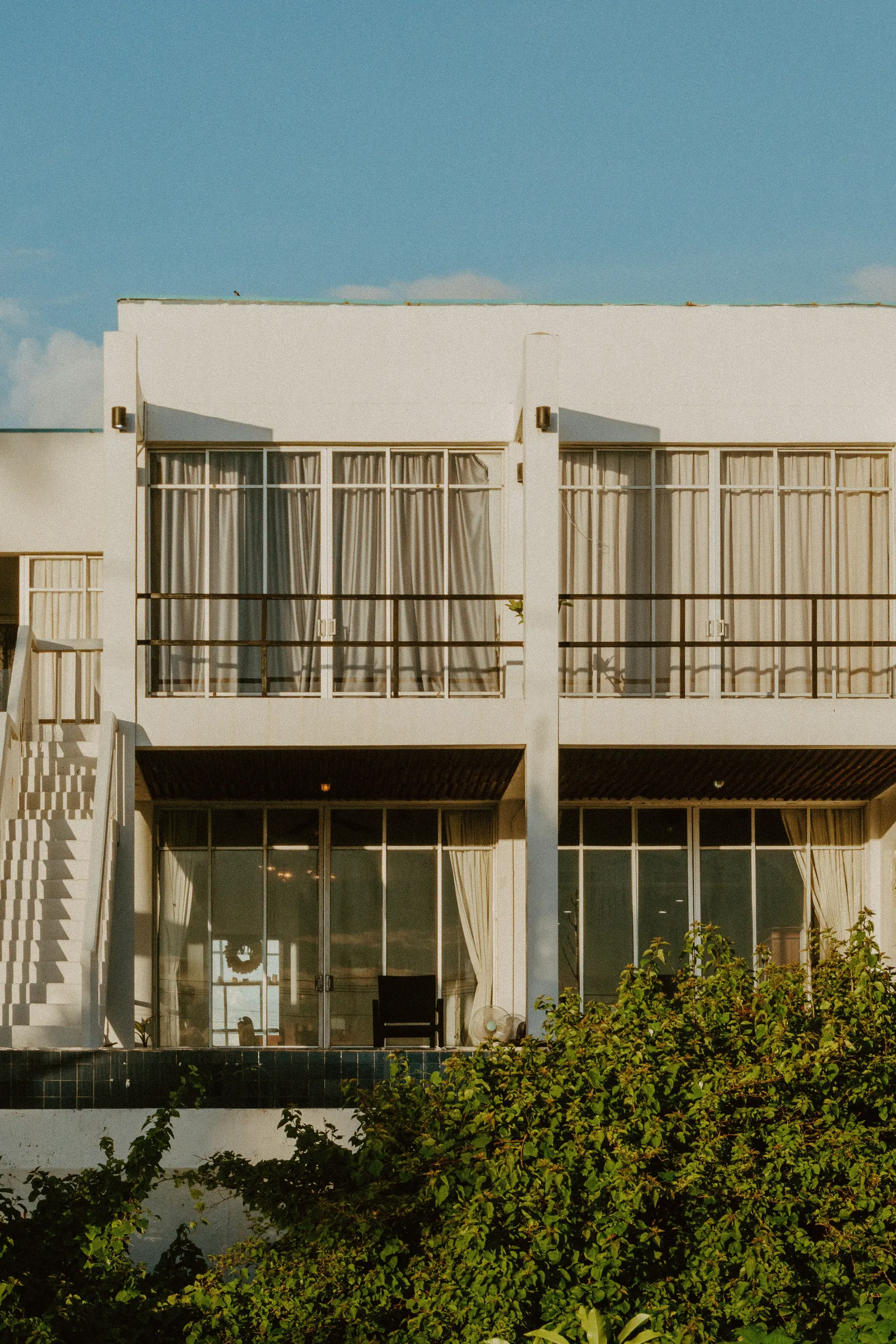 Modern white vacation rental exterior with glass balconies and curtains, framed by greenery
