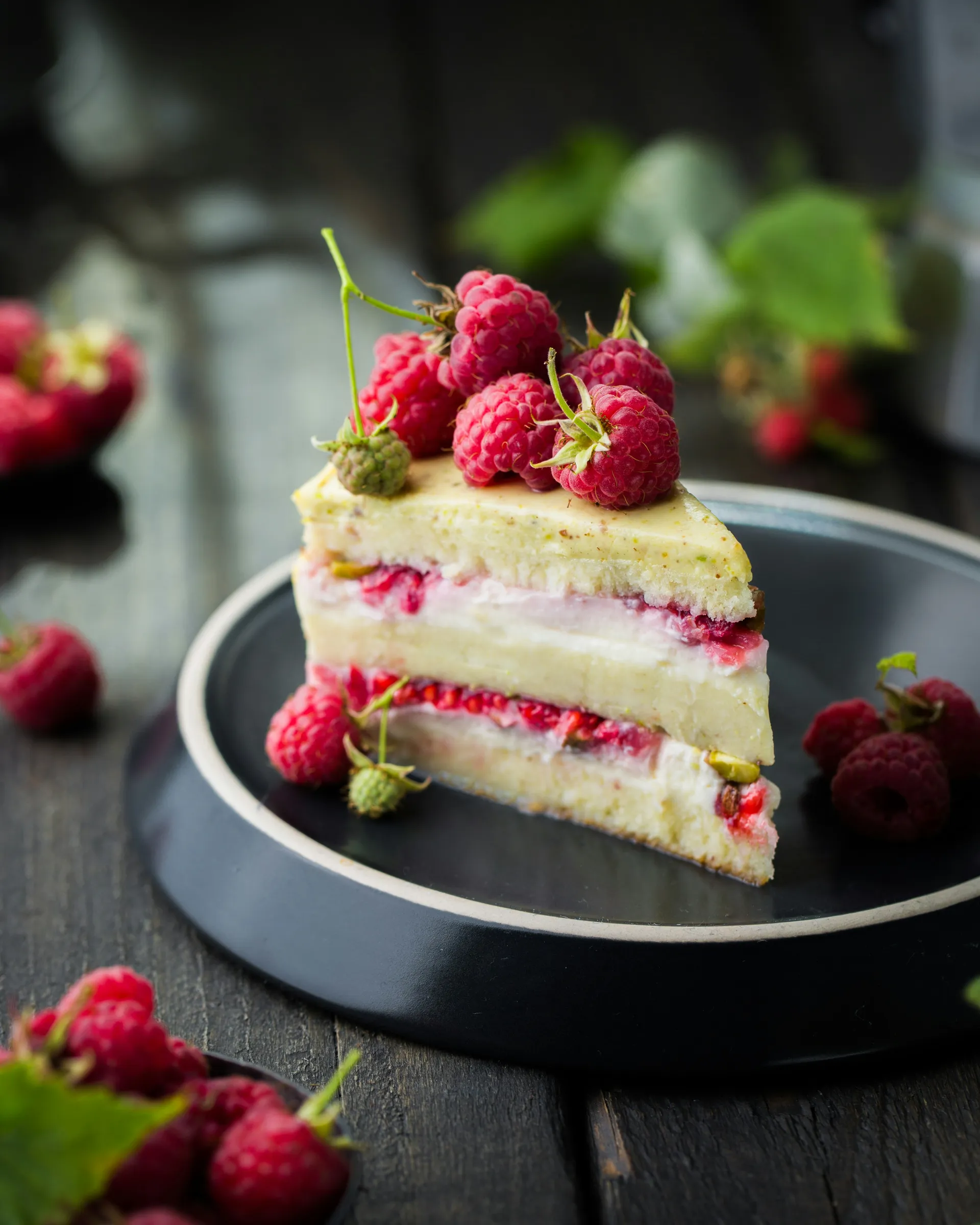 Slice of layered raspberry cream cake topped with fresh raspberries on a dark plate